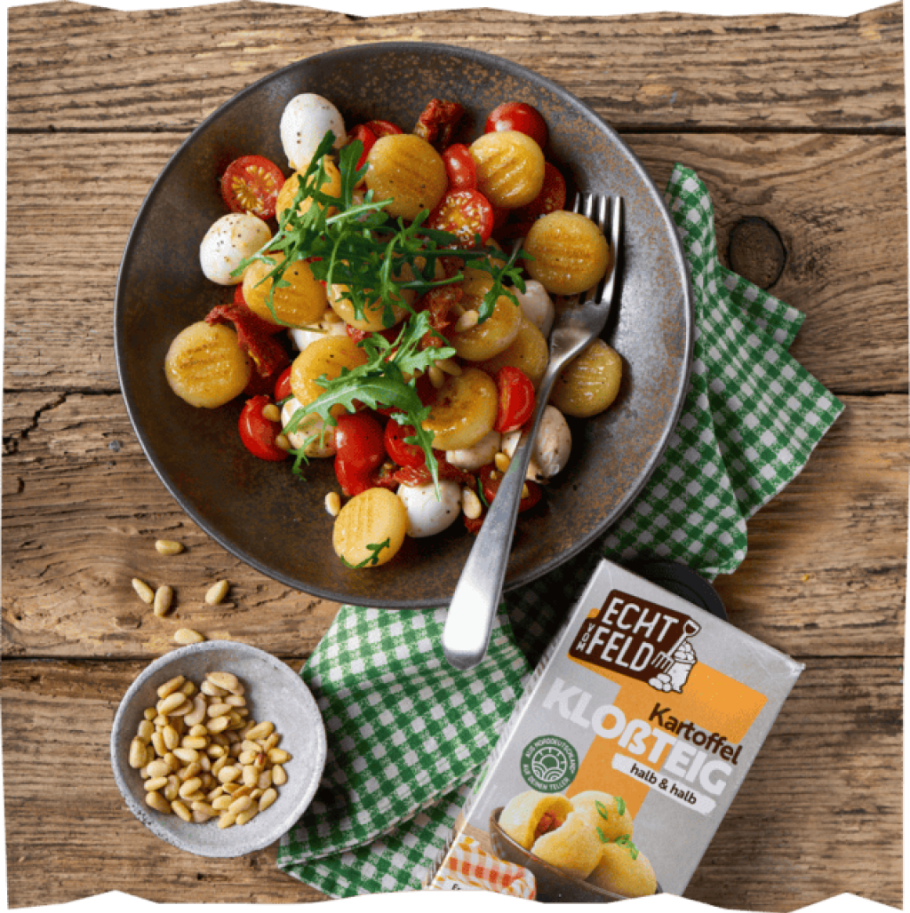 Fertig servierter Gnocchi-Salat mit einer Schale Pinienkernen auf einer grün-weiß karierten Tischdecke mit der Produktverpackung der Echt vom Feld Kloßteig-Mischung.