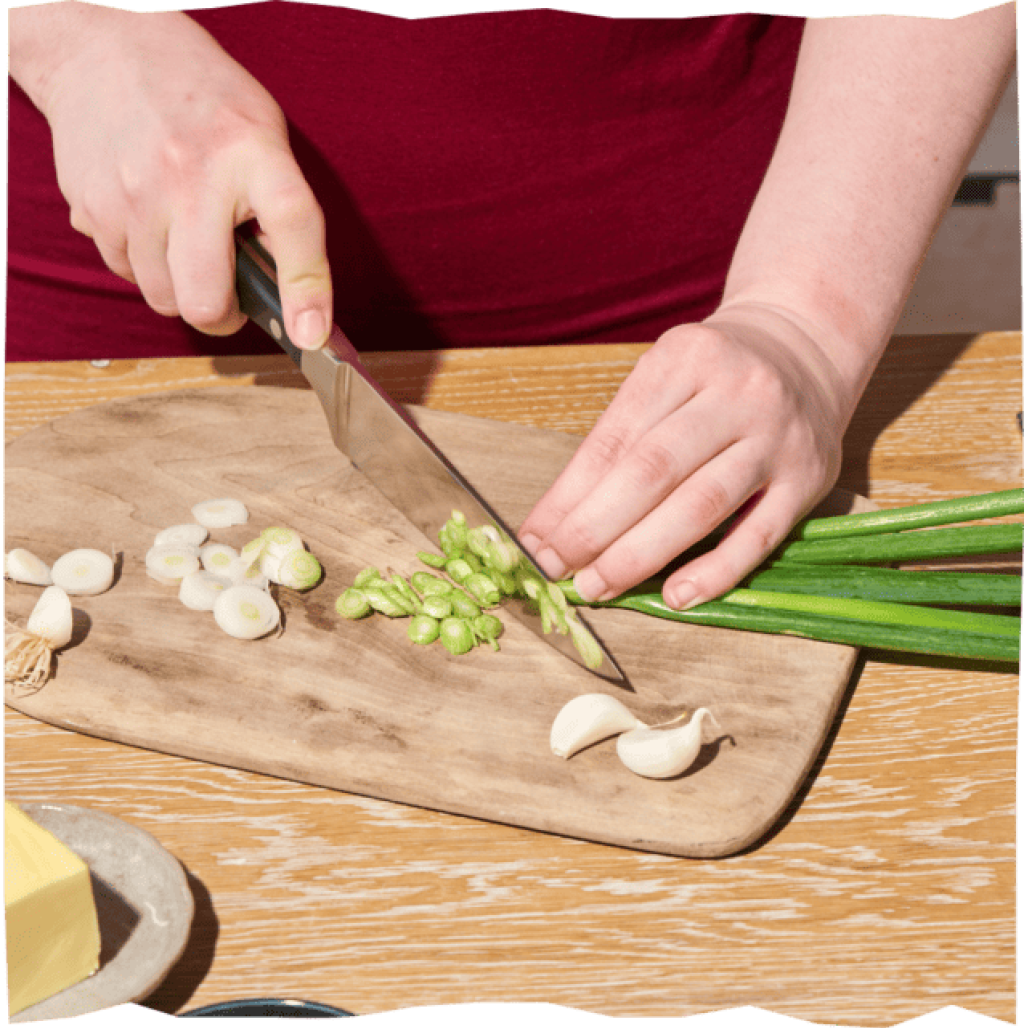 Auf einem Holzbrett werden mit einem Messer die Lauchzwiebeln in Ringe geschnitten. Daneben liegen Knoblauchzehen.