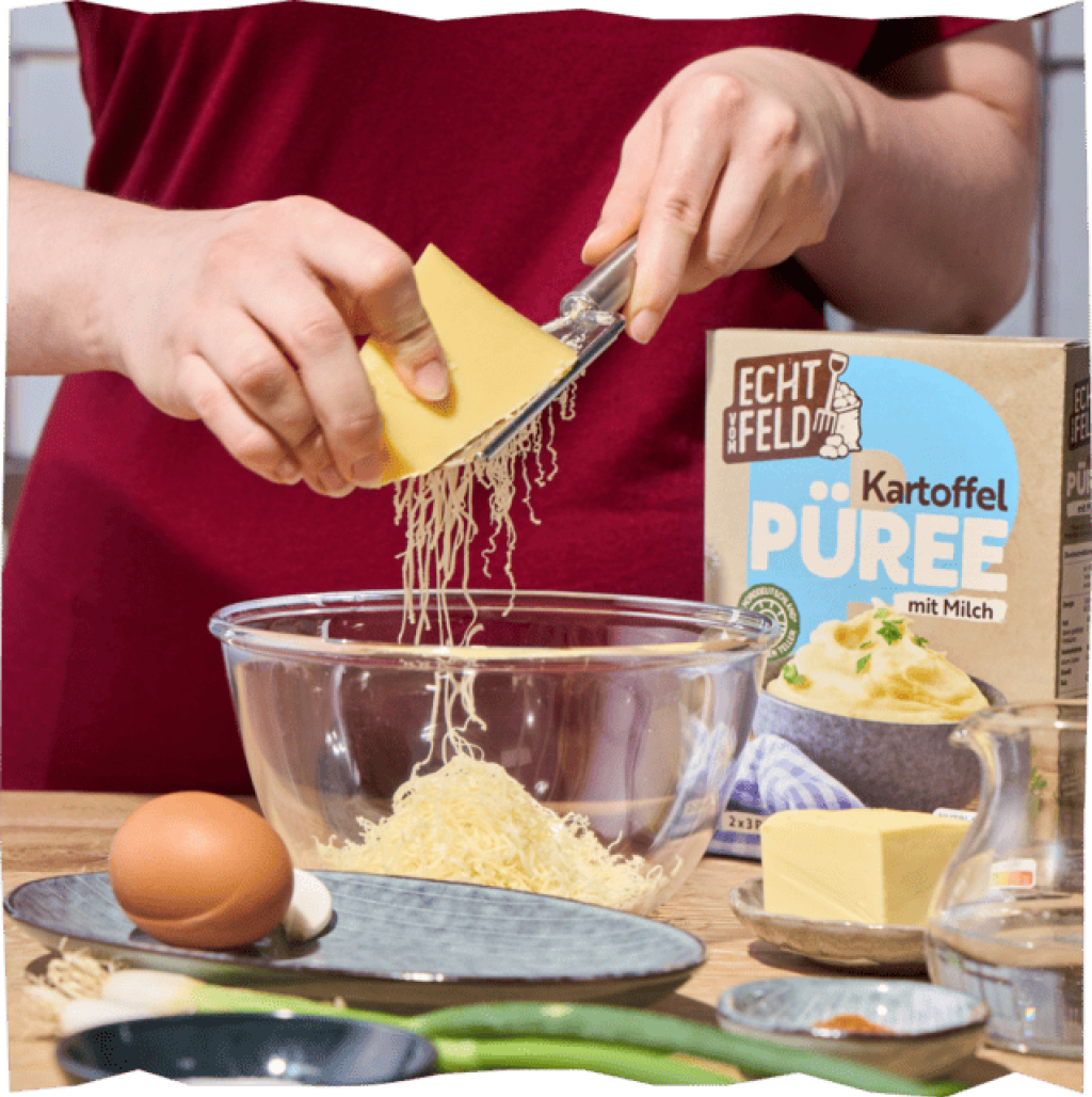 Ein Stück Käse wird mit einer Reibe über eine Glasschüssel
geriebenen. Davor liegen Eier, Lauch und Butter.