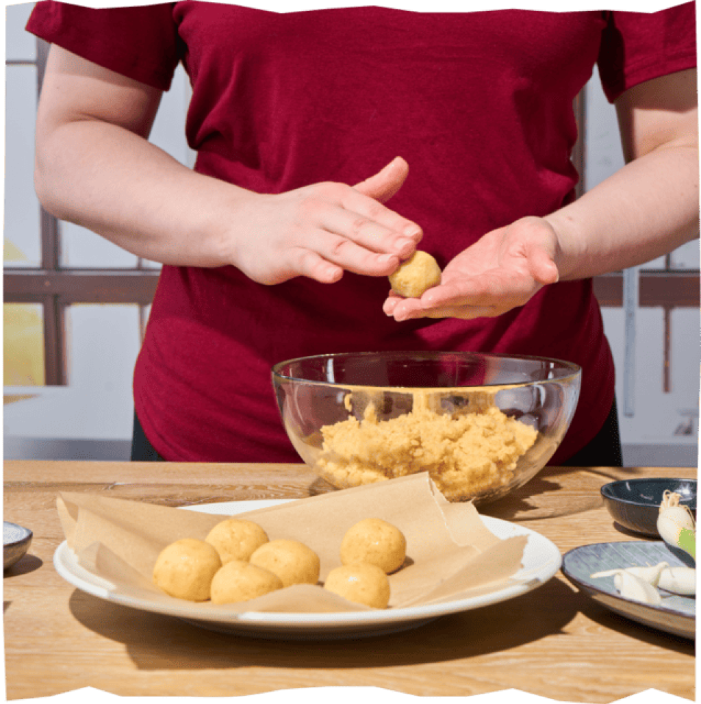Eine Frau im roten Shirt ist im Anschnitt zu sehen, sie rollt aus
dem Kartoffel-Teig kleine Bällchen.
