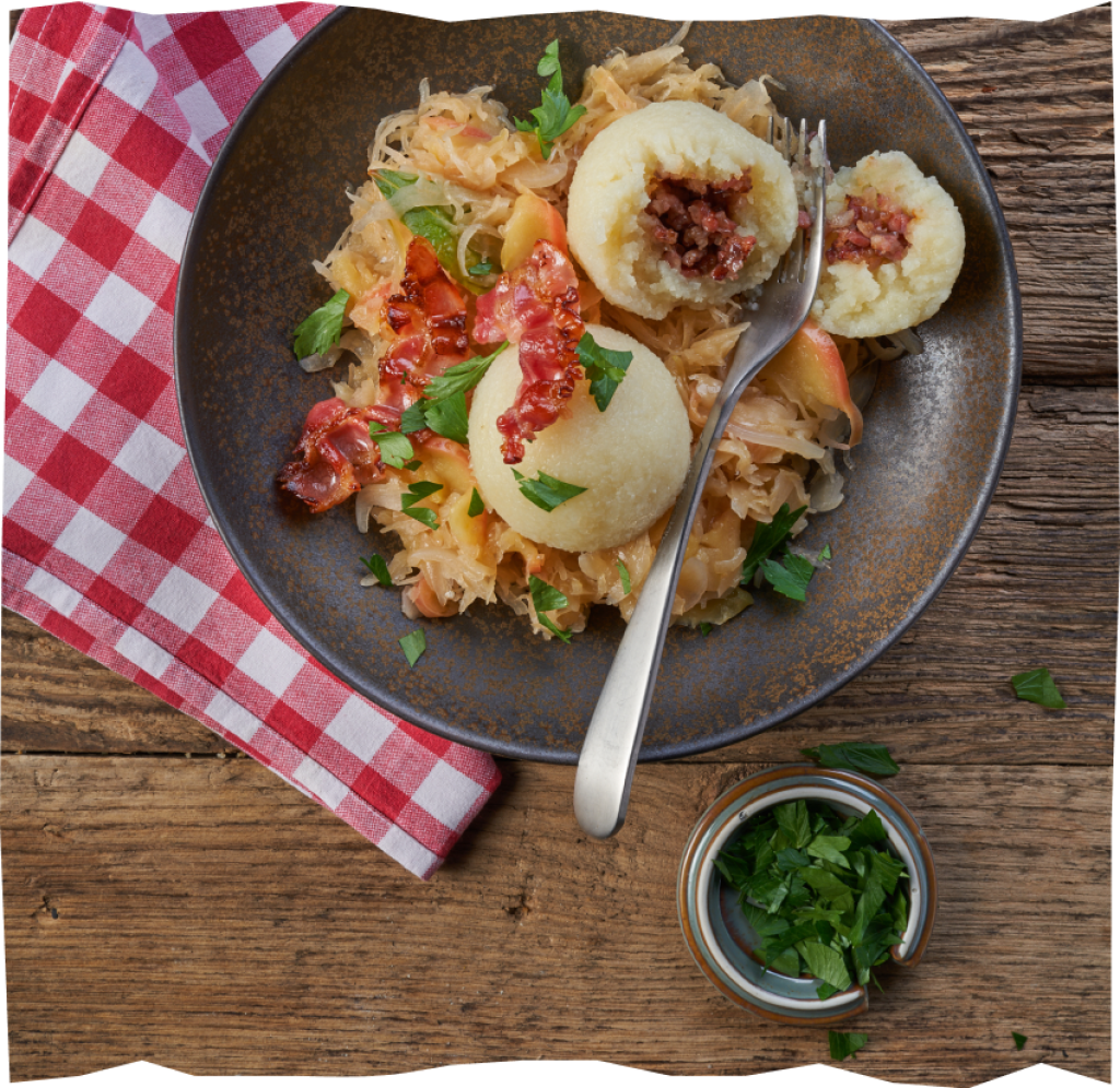 Kartoffelklöße mit Speck, Zwiebeln & Sauerkraut sowie Kräuter-Schälchen auf einem Holztisch serviert.