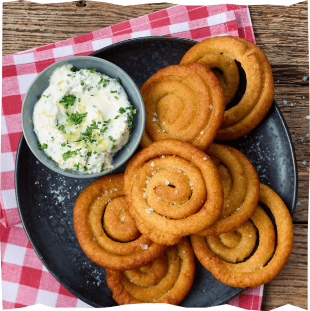 Knusprige Kartoffelswirls auf schwarzem Teller mit rot-weiß karierter Tischdecke und Schale Kräuterdip.