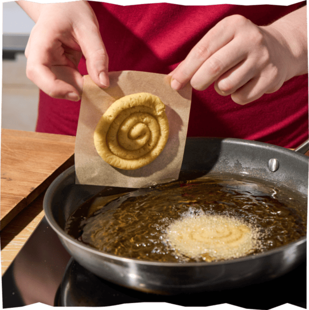 Hände schieben Kartoffel-Swirl vom Backpapier in heißes Öl, ein weiterer Swirl schwimmt und backt in der Pfanne.