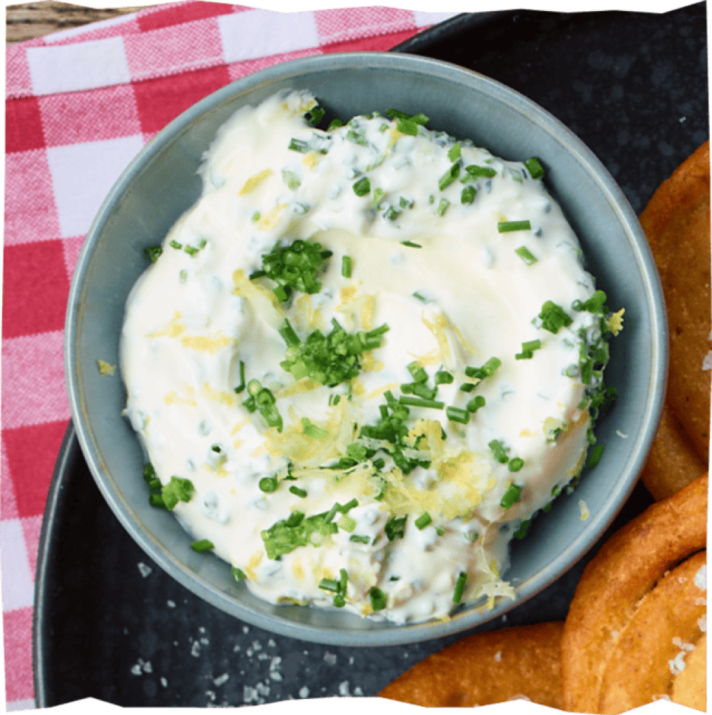 Keramikschale mit cremigem Kräuterdip auf rot-weiß karierter
Tischdecke.