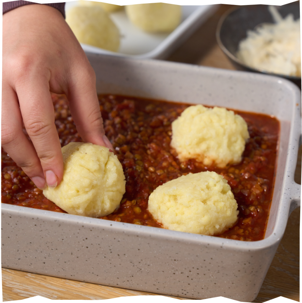 Nach Packungsanweisung verarbeitete Echt vom Feld Kartoffelklöße halb & halb, die in einer Ofenform auf einer Linsen-Bolognese gebettet werden.