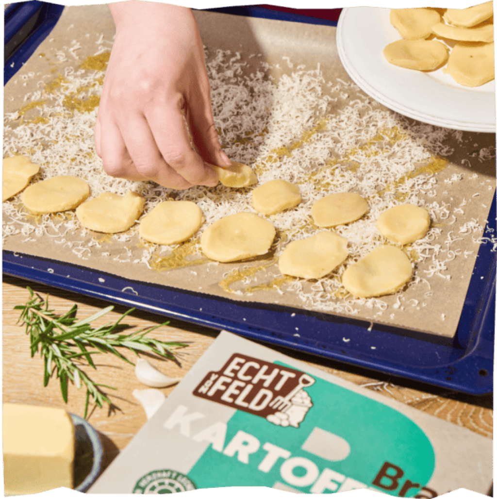 Kartoffelscheiben werden auf geriebenen Käse auf das Backblech gelegt, eine Packung Bratkartoffeln und Rosmarin daneben.