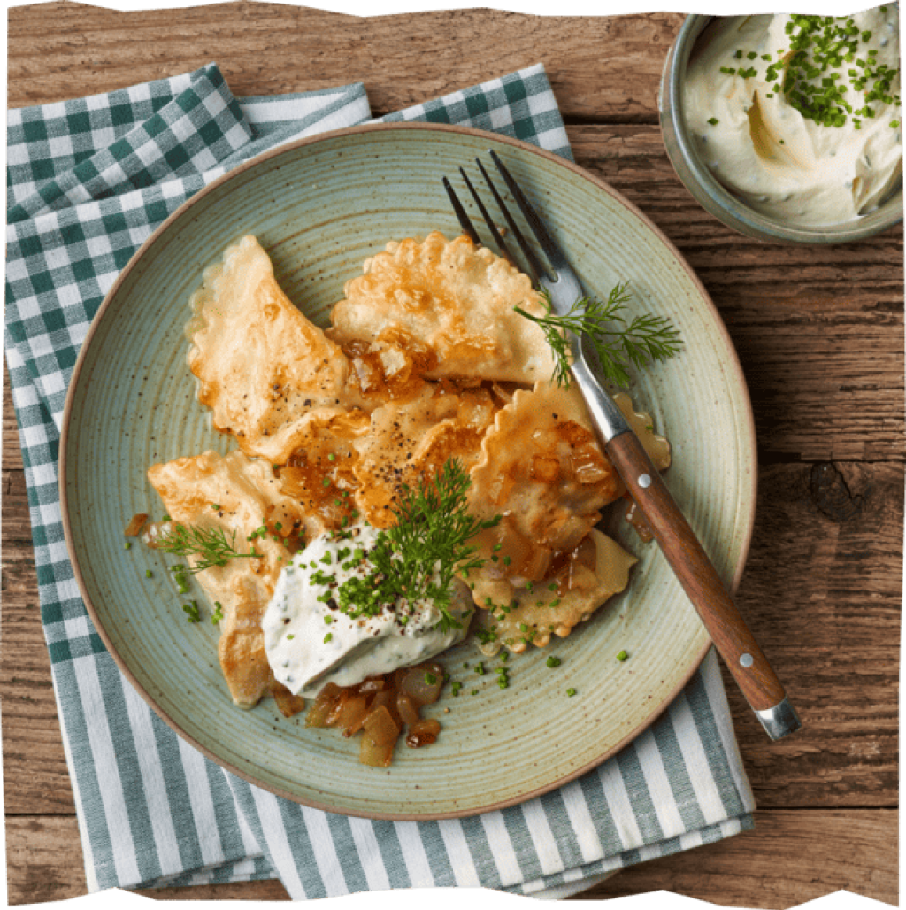 Kartoffel-Pilz-Piroggen mit Schmad und Kräutern auf einem Holztisch und einer grün karierten Stoffserviette.