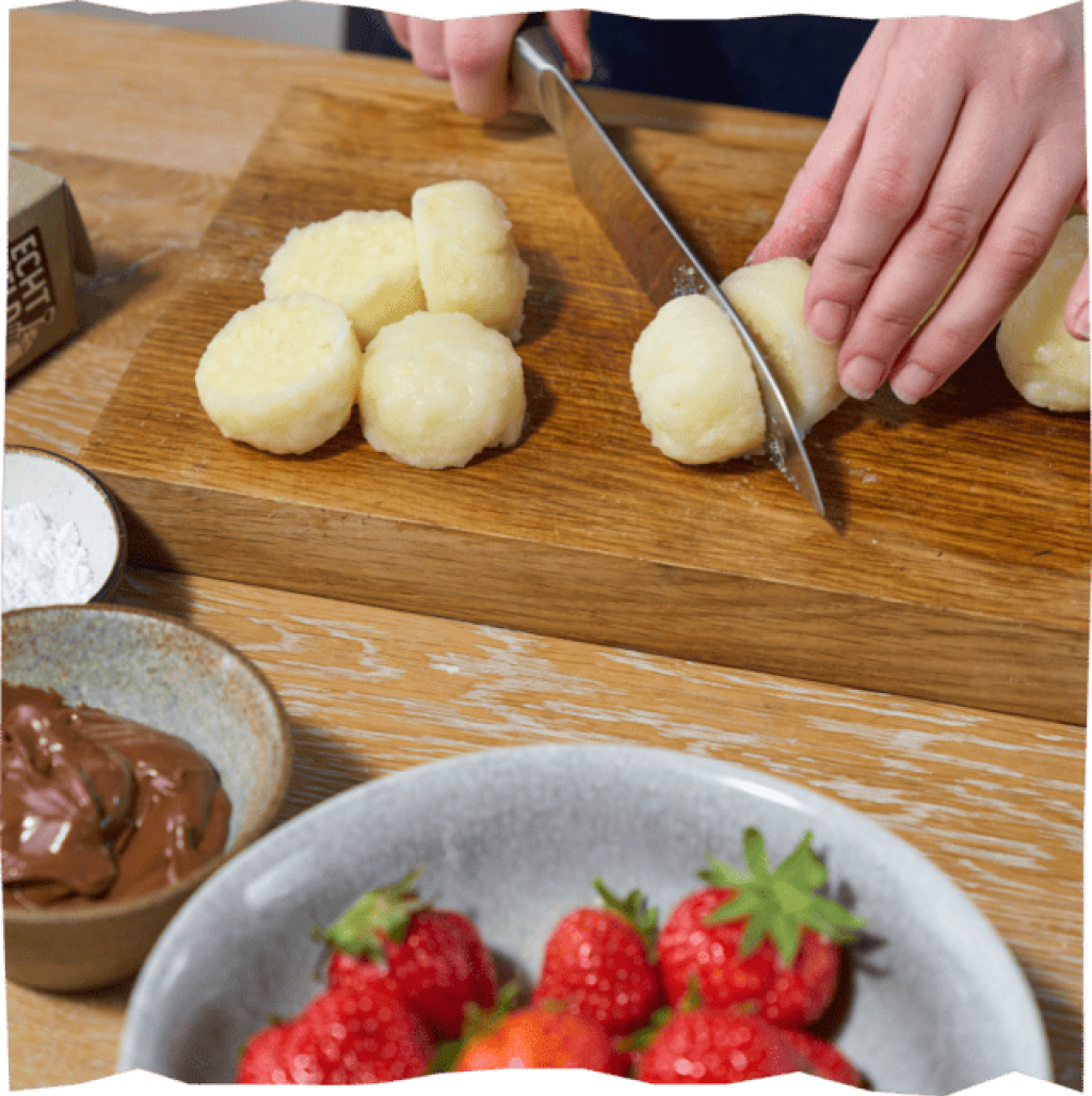 Halbierte Mini-Kartoffelklöße auf einem Holzbrett mit Schokoladensoße und Erdbeeren.